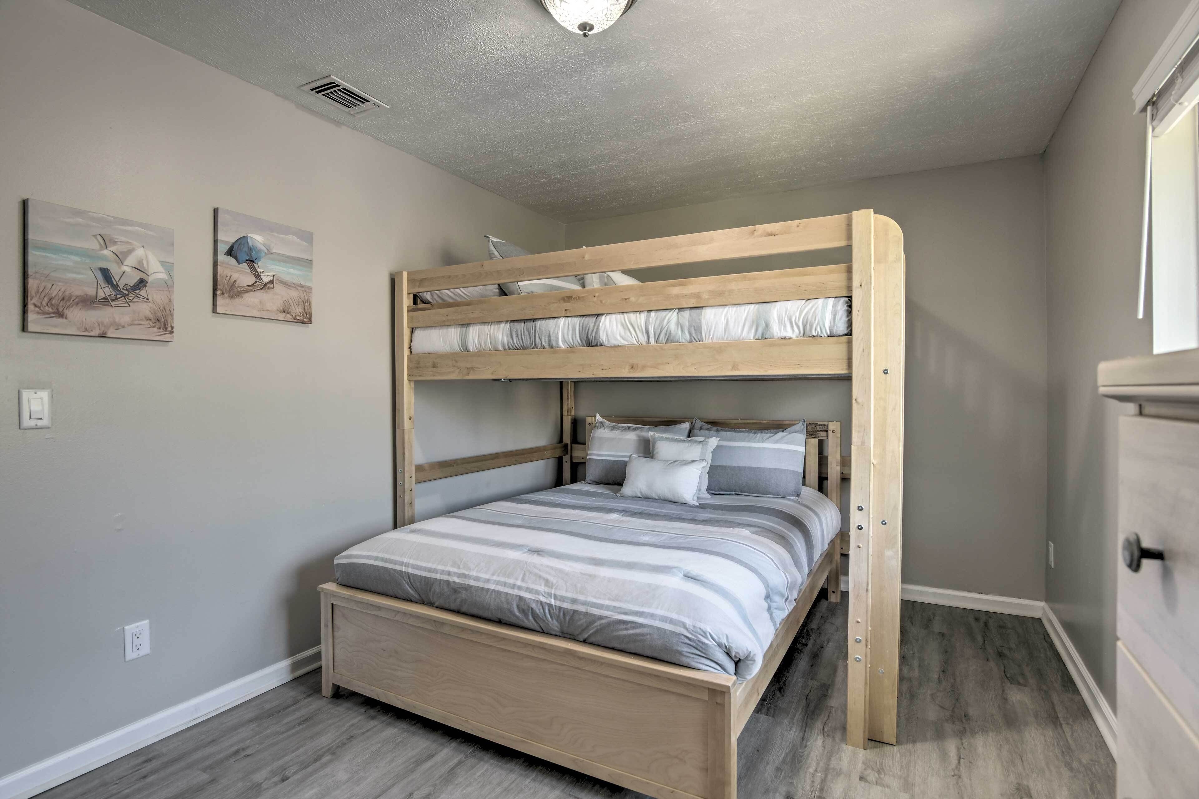 Un letto a castello con struttura in legno e materasso a righe grigio. Ci sono due cuscini sul letto inferiore. La stanza ha pareti grigio chiaro e pavimento grigio. Si vede una finestra sul lato destro dell'immagine.