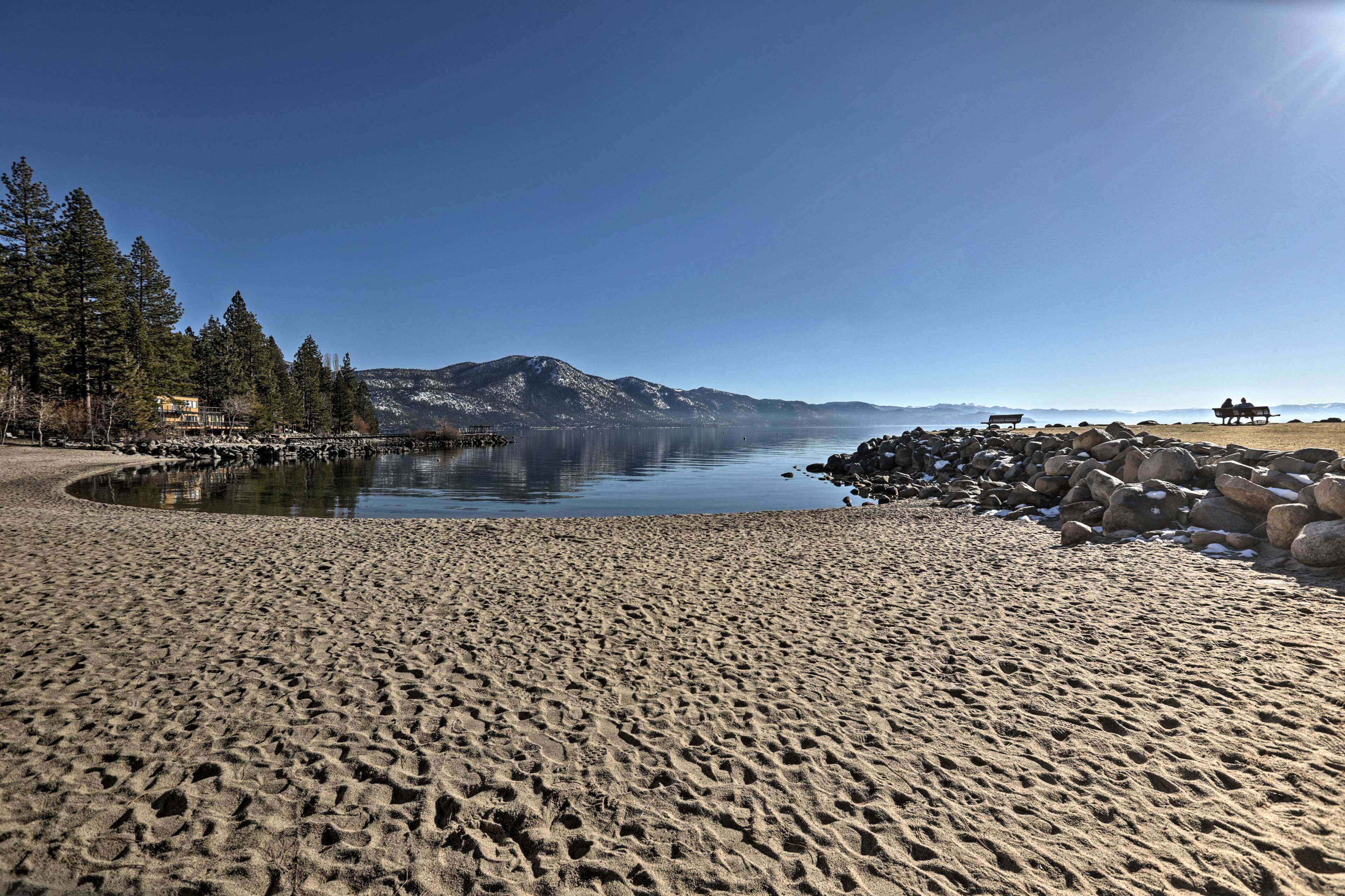 Une plage de sable bordée d’un lac aux eaux calmes, d’une côte rocheuse et, au loin, d’une montagne.