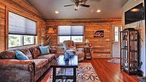 A log cabin interior with a leather couch, a wooden coffee table, a ceiling fan, and a television mounted on the wall.