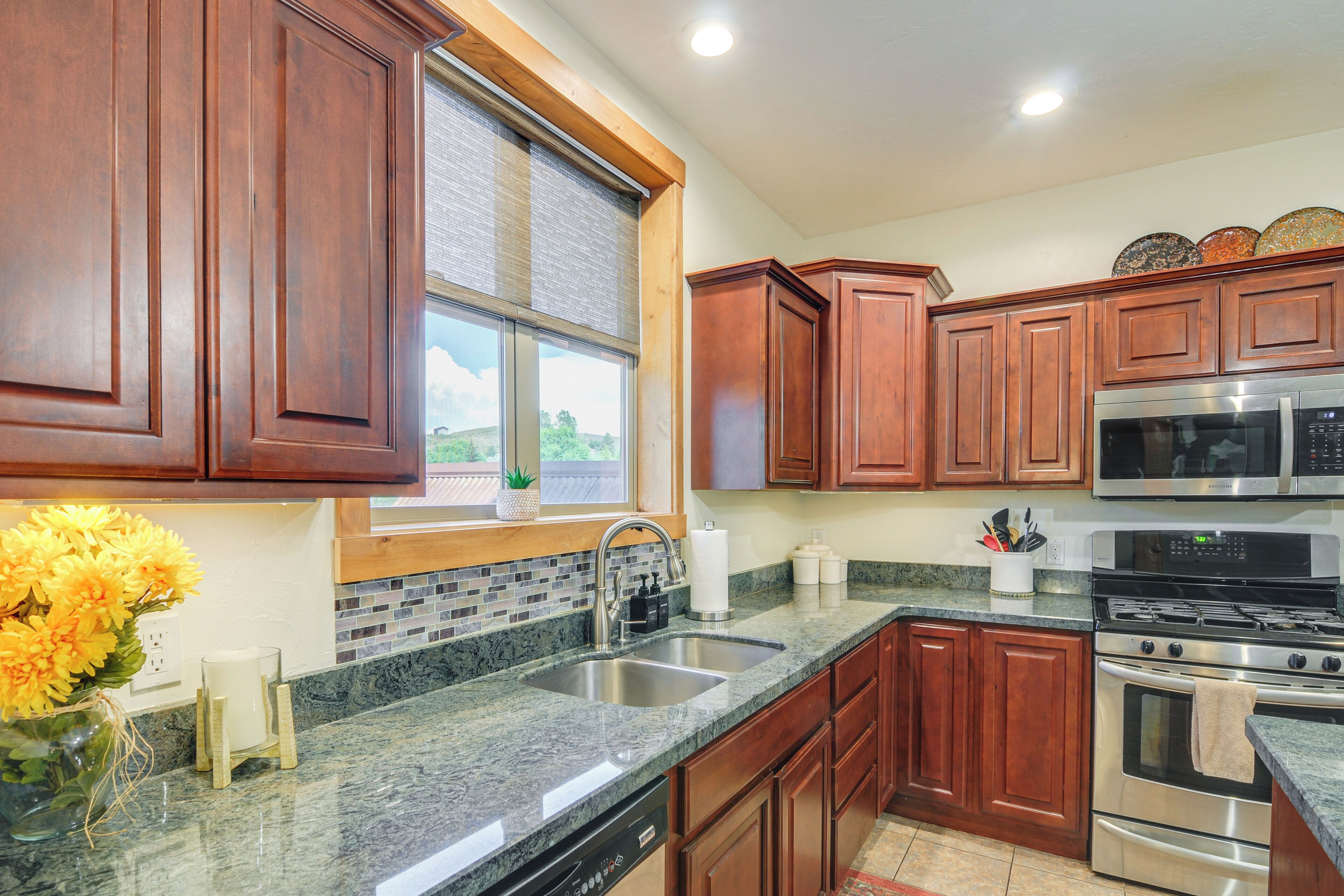 A kitchen with granite countertops, stainless steel appliances, wooden cabinets, a window with blinds, and a vase with flowers on the counter.