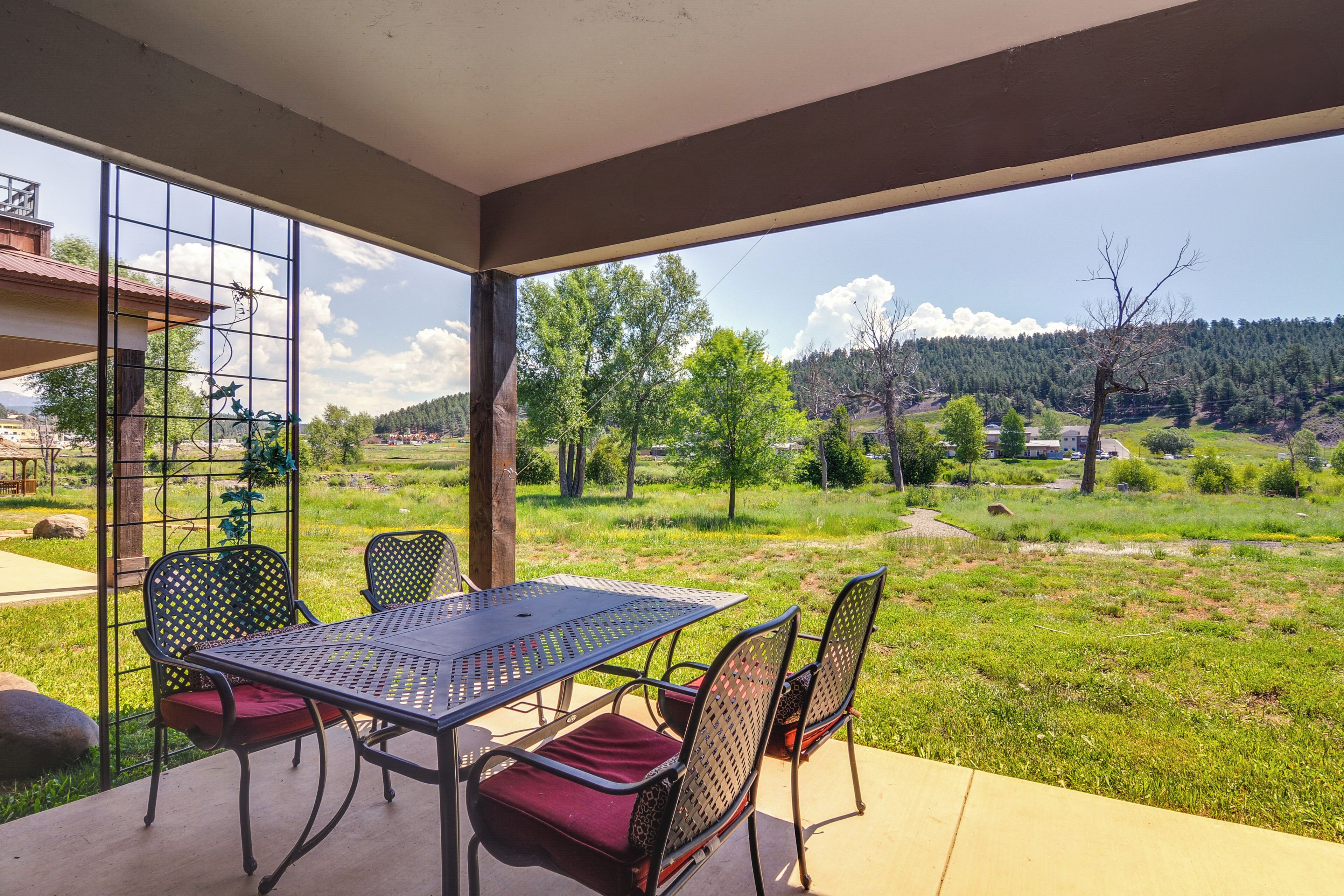 A covered patio with a table and chairs, a view of a green landscape with trees and hills, and a metal fence.