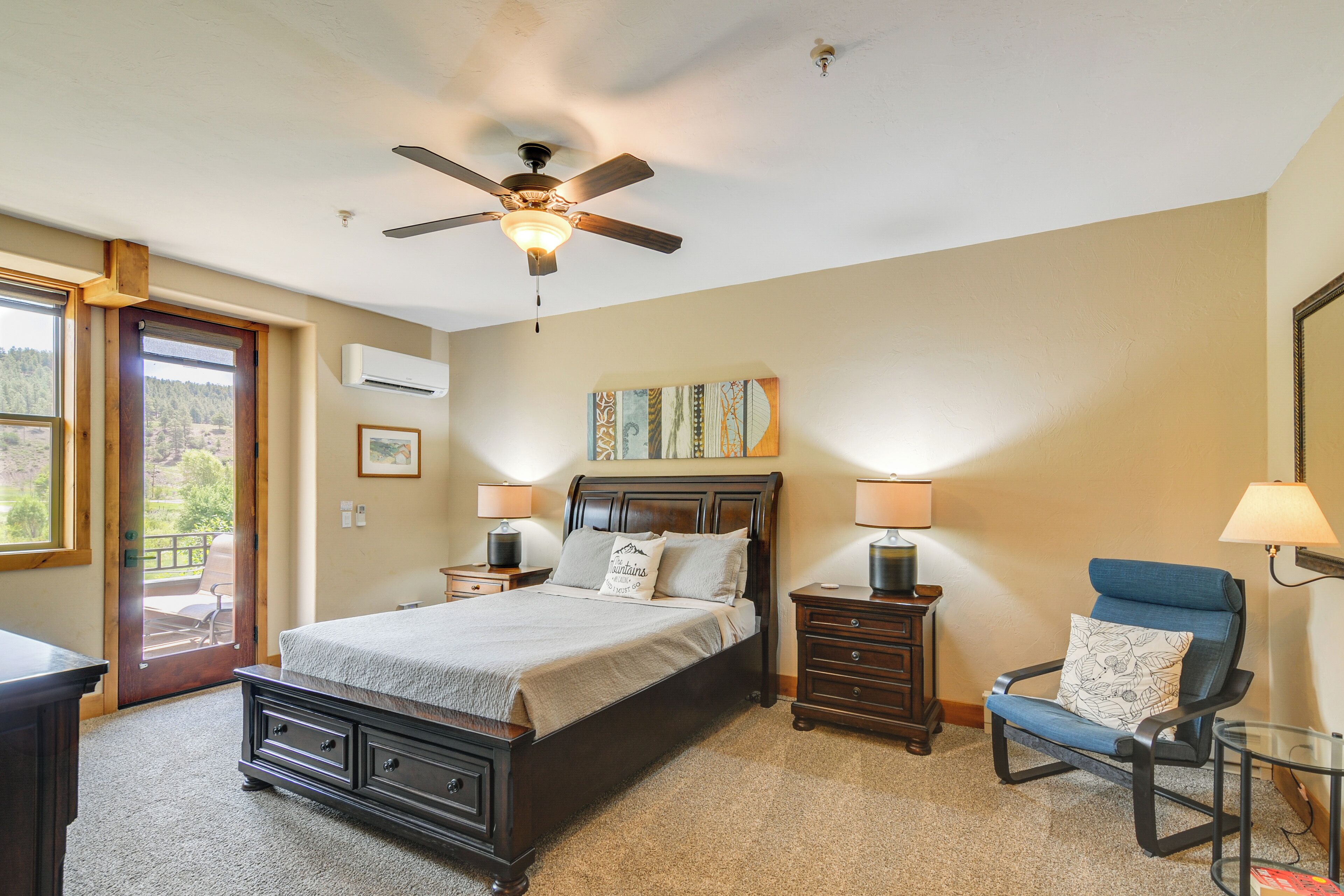 A bedroom with a bed, a ceiling fan, two bedside tables with lamps, a chair, and a door leading to a balcony.