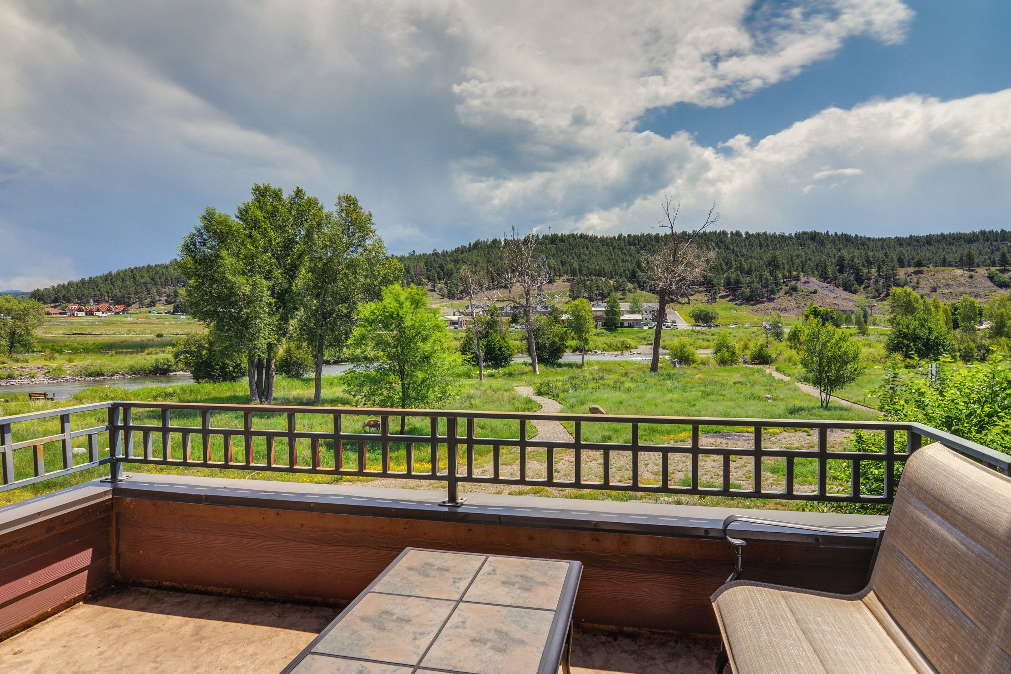 A balcony with a wooden bench and table, overlooking a green landscape with trees and a path.