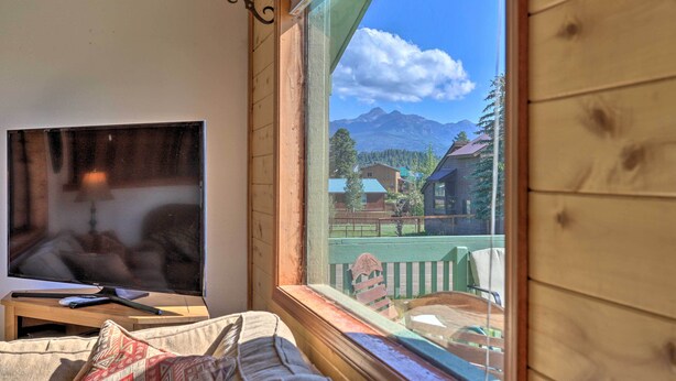 Una habitación con una televisión sobre un soporte de madera, una ventana con vistas a montañas y casas, y una pared de madera.