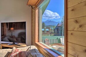 Una habitación con una televisión sobre un soporte de madera, una ventana con vistas a montañas y casas, y una pared de madera.