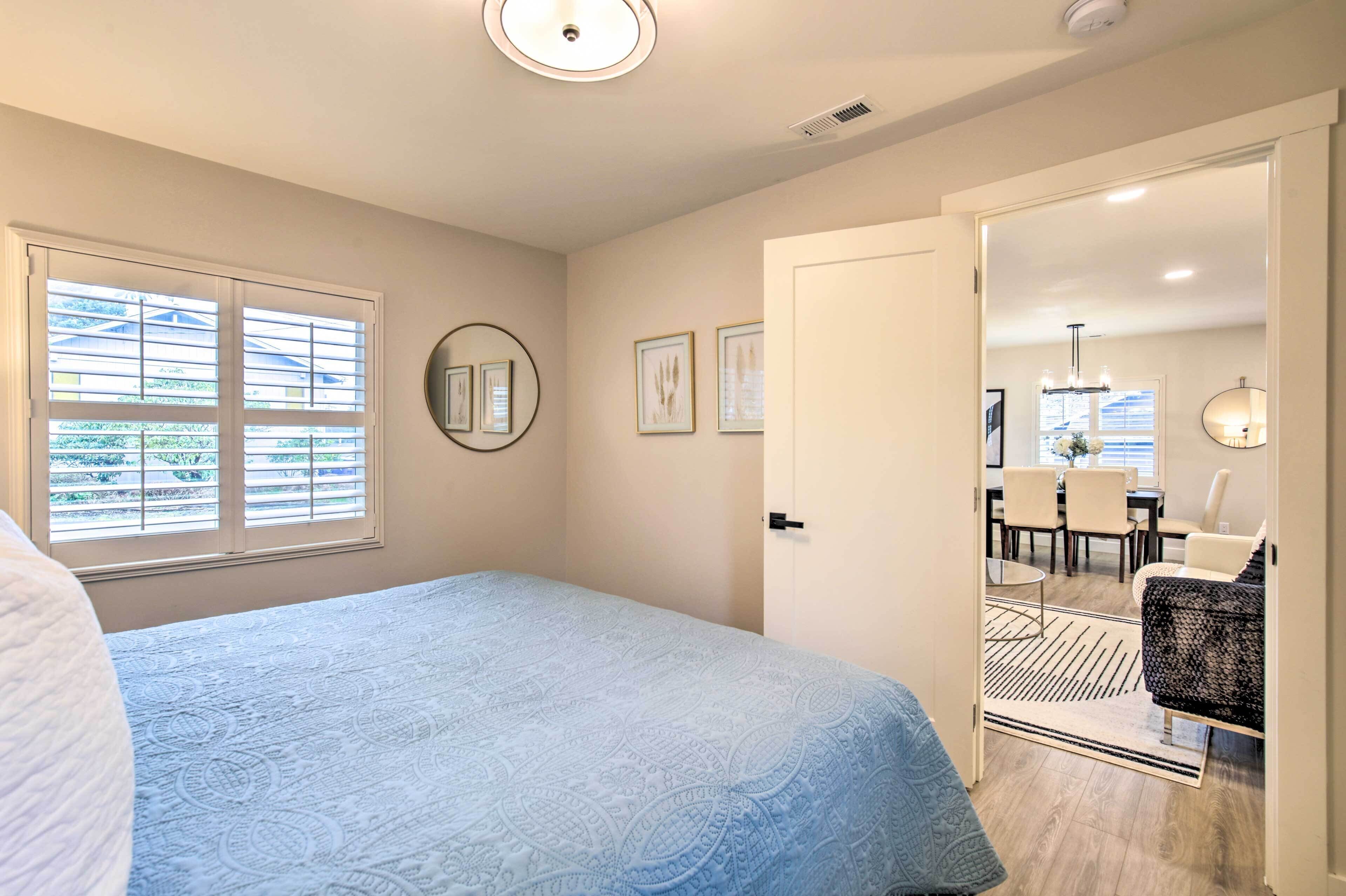 A bedroom with a bed, a door leading to a dining area, and a window with blinds.
