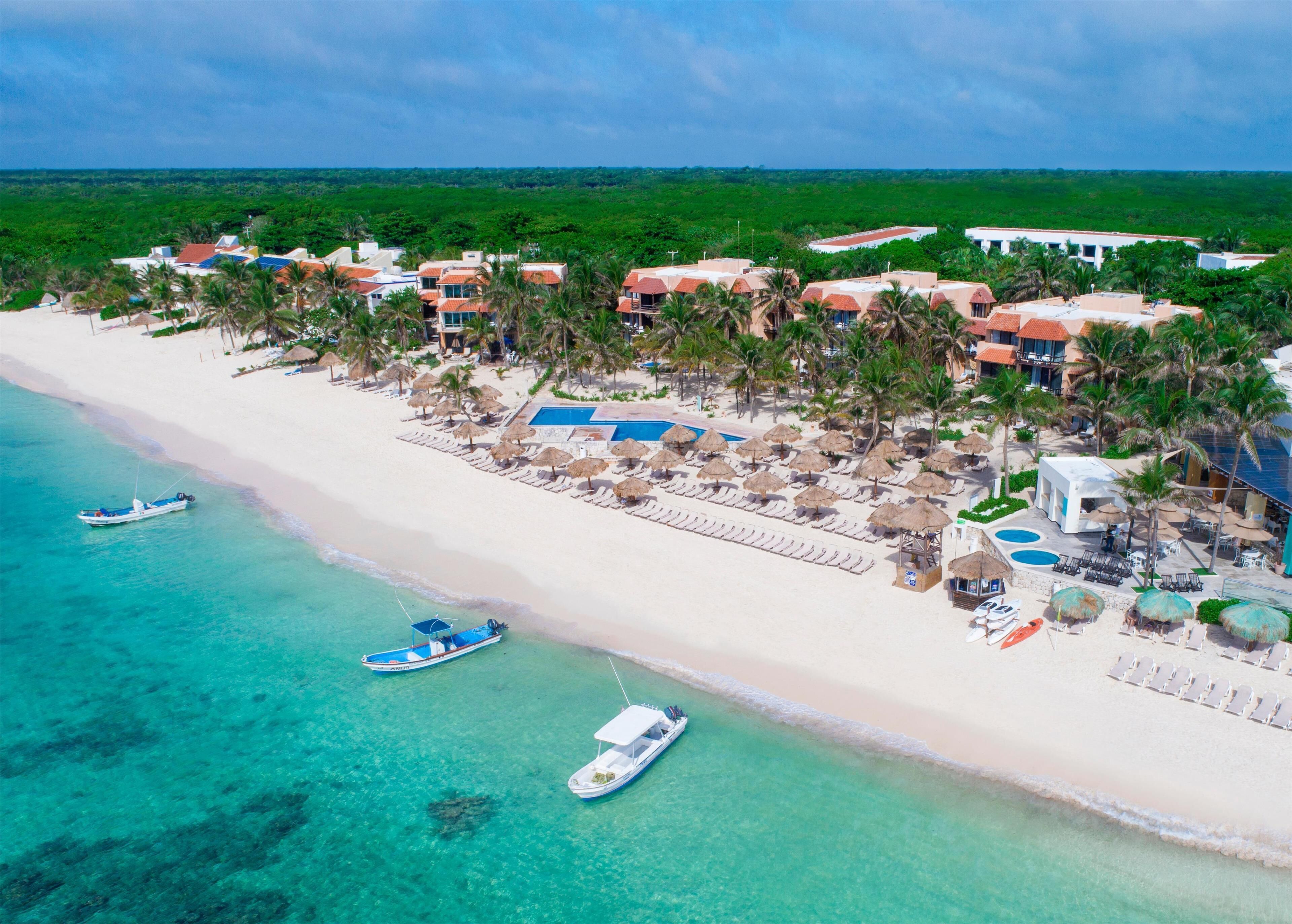 An aerial view of a beach resort with multiple buildings, a sandy beach, clear turquoise waters, and several boats.