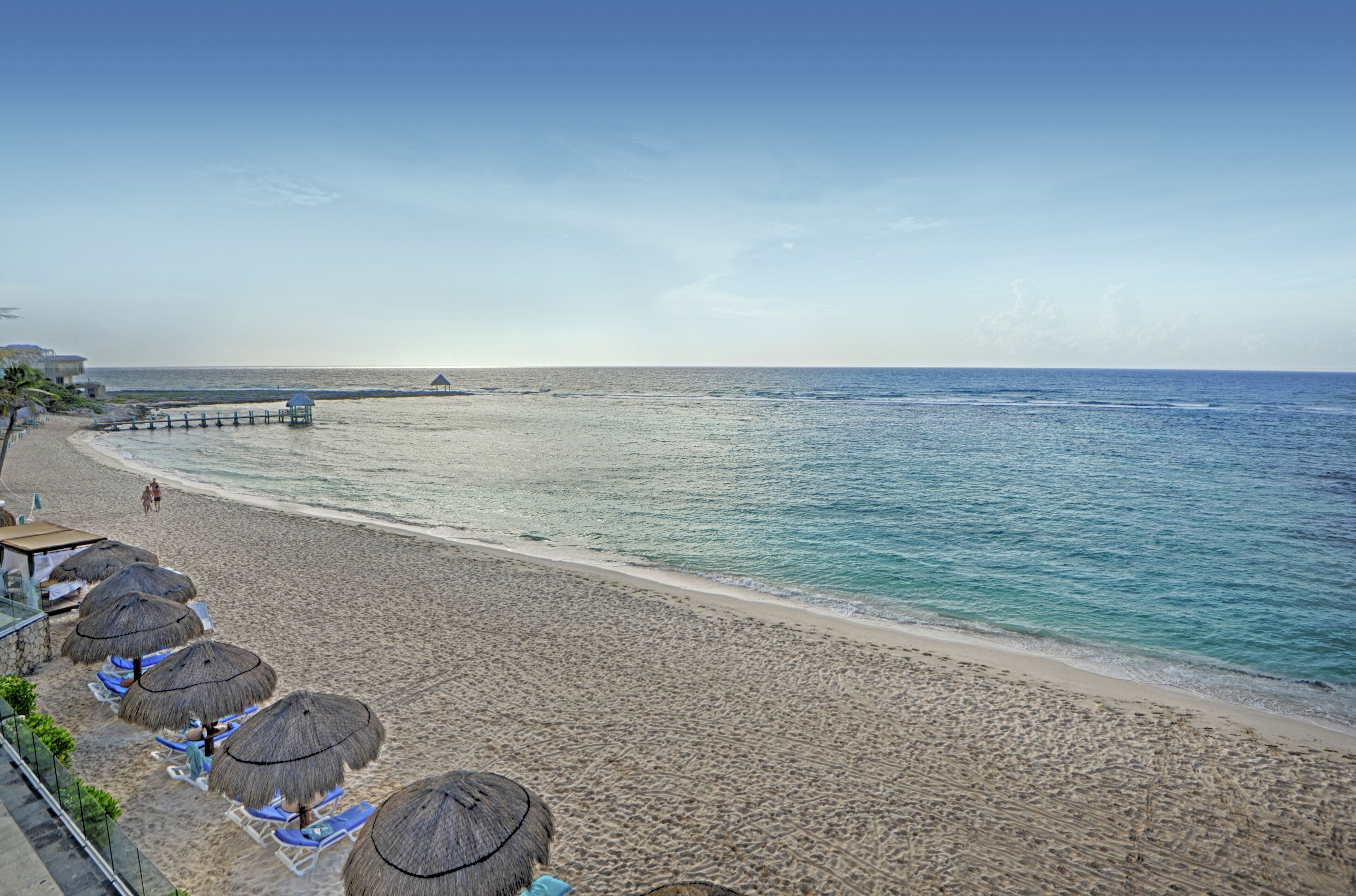 Una spiaggia con ombrelloni di paglia e lettini, un molo che si protende sull'acqua e un cielo sereno.