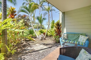 A covered outdoor seating area with a wicker sofa and a small table, surrounded by tropical plants and palm trees. - Oceanfront Close to Ironman Start at Kona Islander (Kailua-Kona)
