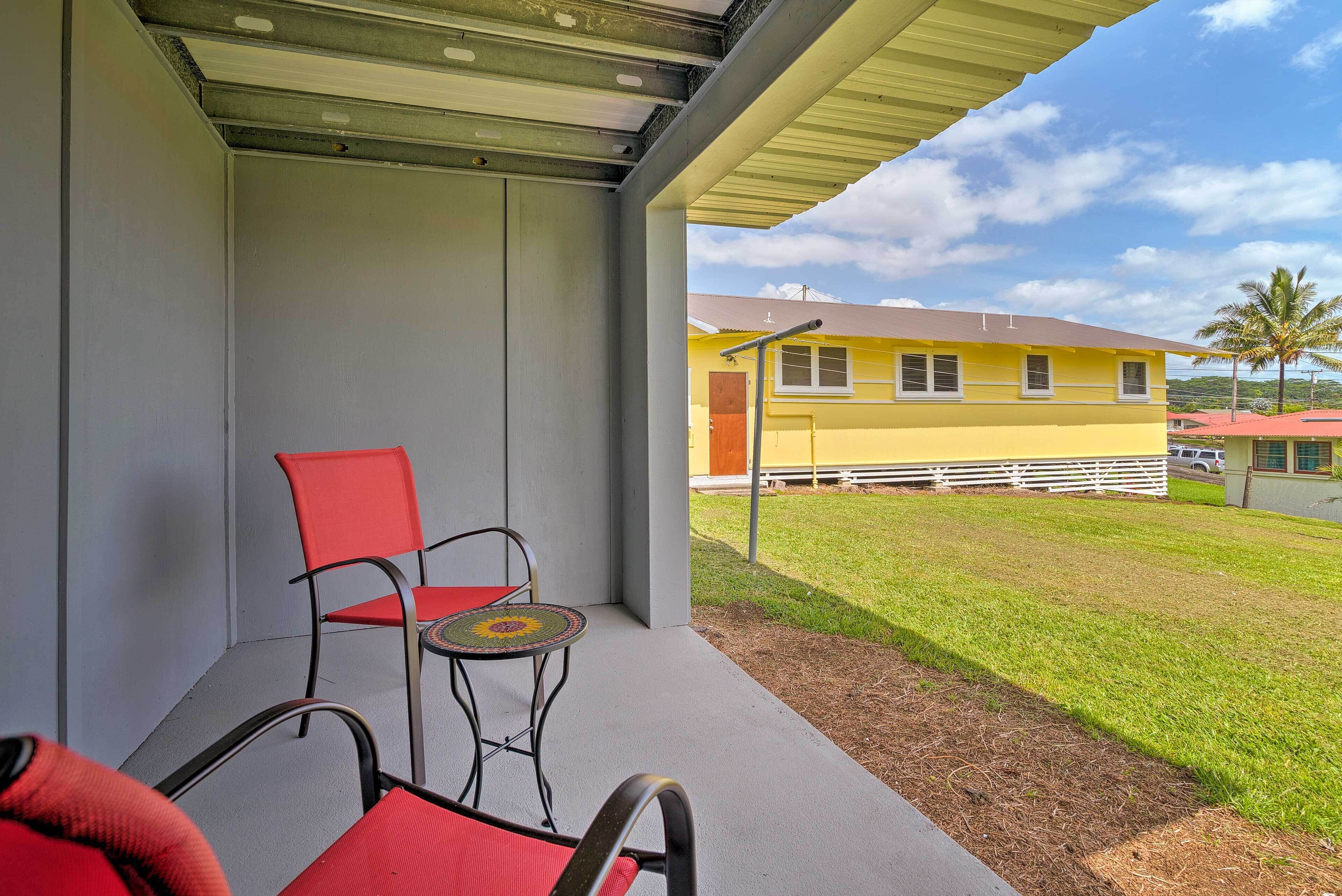 Un espace extérieur abrité avec une chaise rouge et une petite table, un bâtiment jaune en arrière-plan et un ciel dégagé.