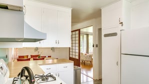 A kitchen with white cabinets, a gas stove, and a refrigerator. There is a dining table with chairs and a view of a traditional Japanese room in the background. - Charming Historic Hilo House Minutes to Beach! (Hilo)
