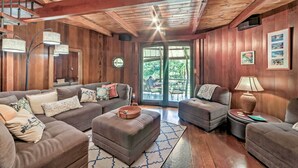 A cozy living room with wooden walls and ceiling, furnished with sofas and a coffee table, and a large window offering a view of the outdoors. - Tropical Cabana w/ Deck, Hot Tub & Lush Scenery! (Keaau)