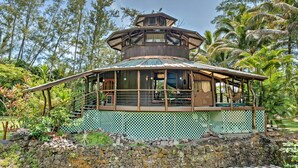 A multi-level wooden structure with a covered deck, surrounded by lush vegetation and a stone wall. - Tropical Cabana w/ Deck, Hot Tub & Lush Scenery! (Keaau)