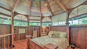 A wooden bedroom with a bed, a nightstand, a lamp, and a chair. - Tropical Cabana w/ Deck, Hot Tub & Lush Scenery! (Keaau)