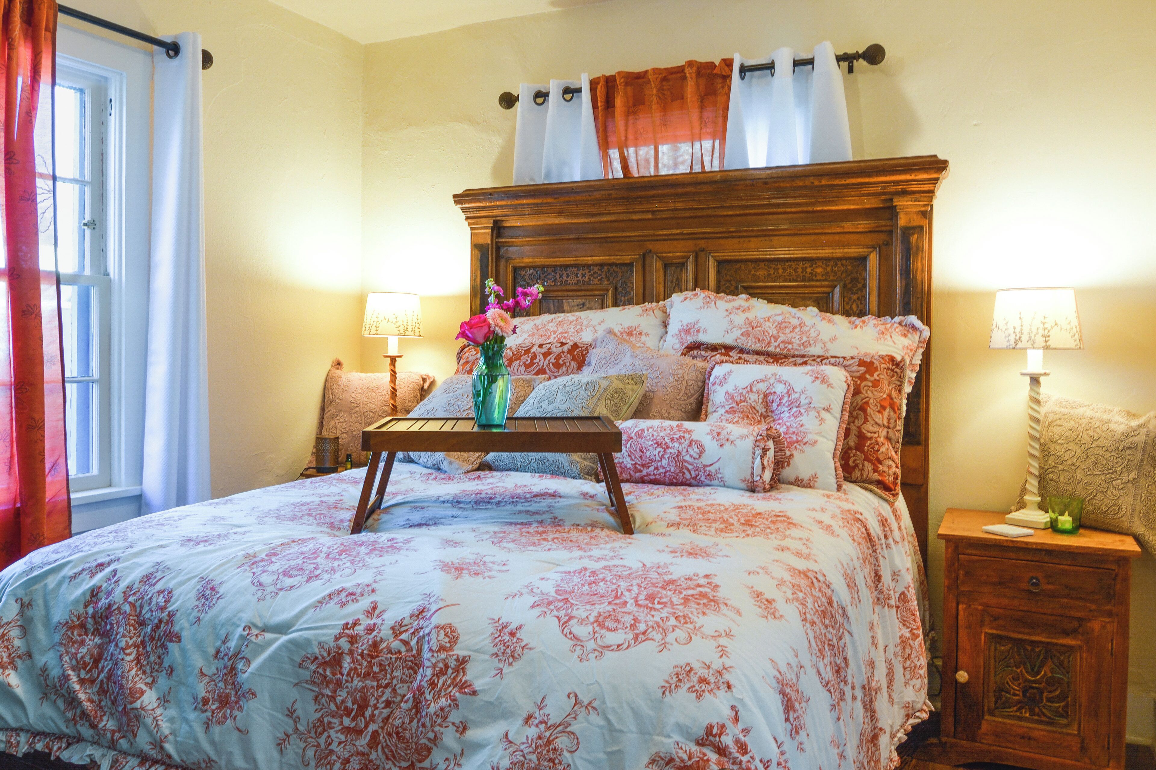 A bedroom with a large bed, floral bedding, a wooden headboard, bedside tables with lamps, and a window with curtains.