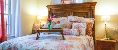 A bedroom with a large bed, floral bedding, a wooden headboard, bedside tables with lamps, and a window with curtains.