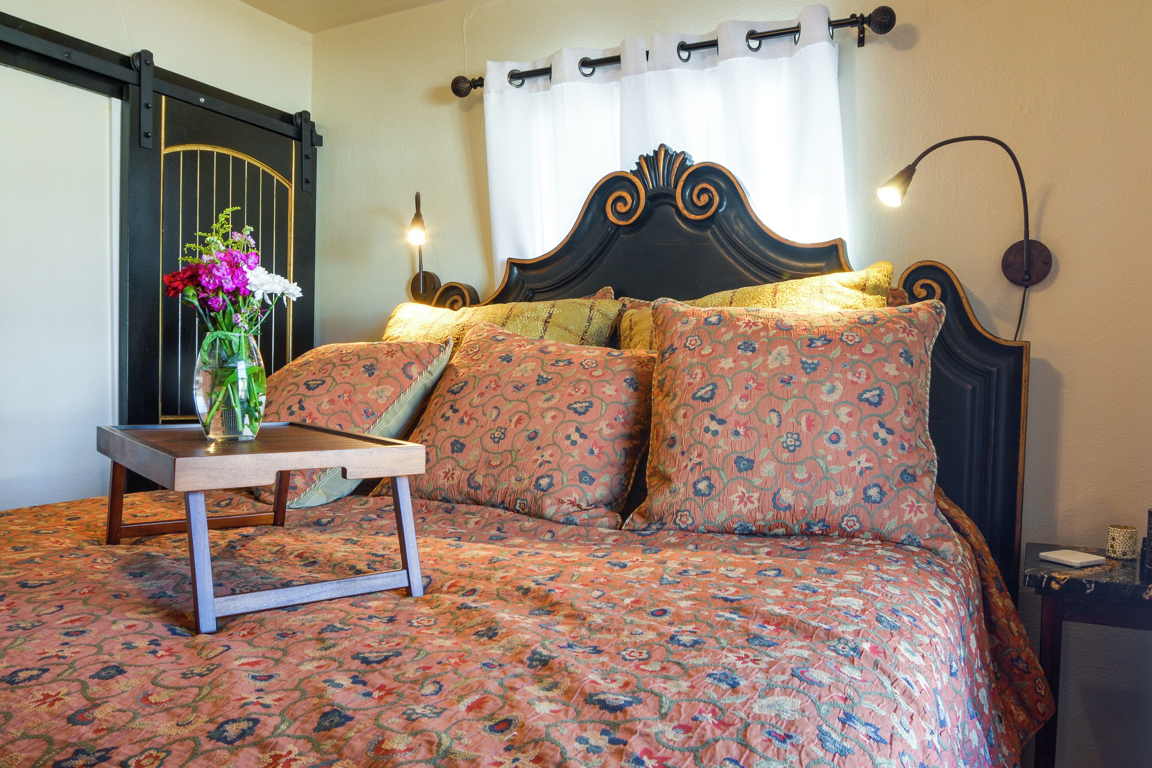 A bed with a floral bedspread, a bedside table with a vase of flowers, and a wall-mounted lamp.