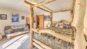 Un dormitorio rústico con una cama con dosel de madera natural, un sofá con cojines estampados y vistas a un paisaje montañoso a través de una puerta abierta.
