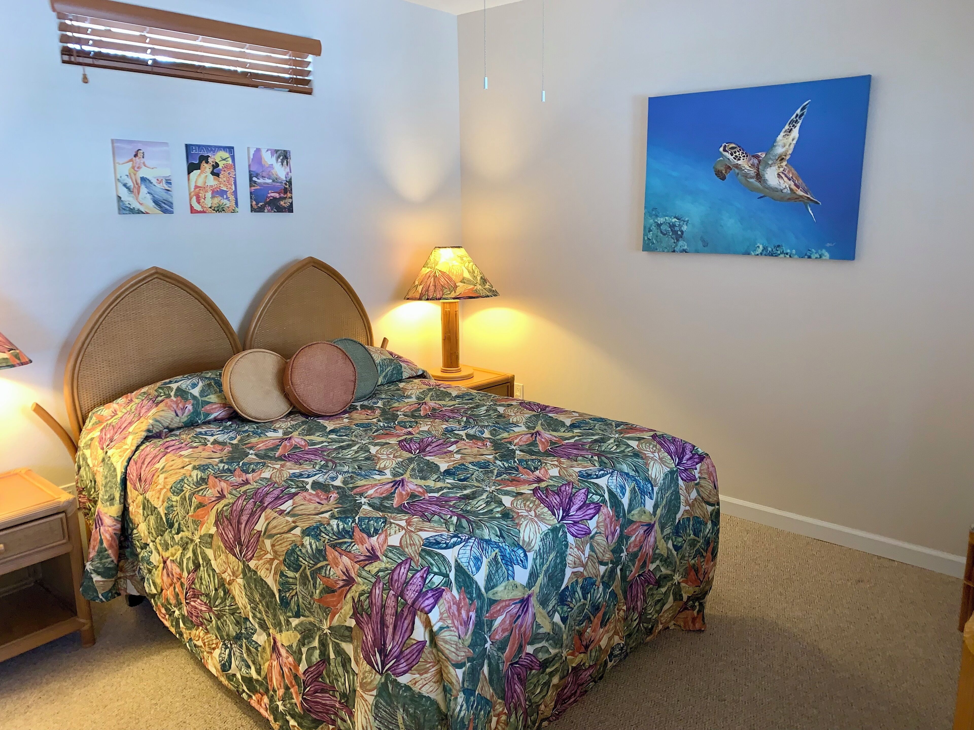 A bedroom with a floral bedspread, a wooden headboard, a nightstand with a lamp, and a wall with framed pictures.