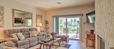 A living room with a sofa, armchairs, a coffee table, and a fireplace. There is a television mounted on the wall and a window with a view of the outdoors.