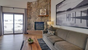 A living room with a stone wall, a fireplace, a couch, and a large window with a view of a snowy landscape. - Balcony & Ski Slope Views: Idyllic Granby Condo! (Granby)
