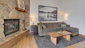 A living room with a stone fireplace, a gray sofa, a wooden coffee table, and a framed landscape picture on the wall. - Balcony & Ski Slope Views: Idyllic Granby Condo! (Granby)
