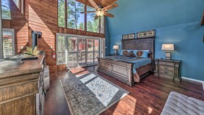 A bedroom with a wooden ceiling, a large bed, a nightstand, and a window with a view of trees. - Hot Tub, Game Room & Sauna: Ruidoso Hideaway! (Ruidoso)