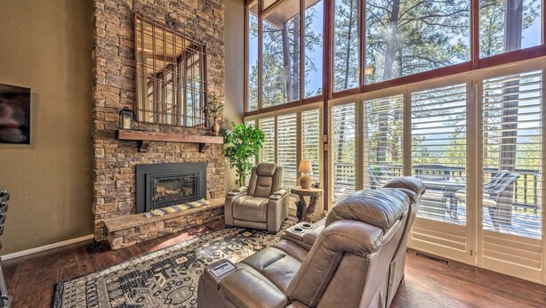 A spacious living room with a stone fireplace, leather recliner, and large windows offering a view of trees. - Hot Tub, Game Room & Sauna: Ruidoso Hideaway! (Ruidoso)