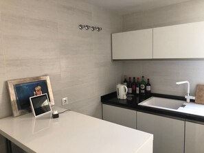 Une cuisine moderne avec un plan de travail blanc, un évier et des placards. On peut y voir des bouteilles de vin et une bouilloire posées sur le plan de travail.