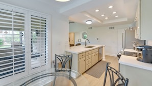 A modern kitchen with a central island, white cabinetry, and a large window with blinds. - Quiet Palm Desert Condo: Workspace & Pool Access! (Palm Desert)