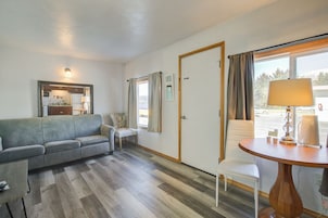 Una habitación con un sofá gris, una mesita con una lámpara, una silla, suelo de madera, una puerta y una ventana con cortinas.