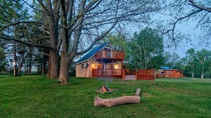 Uma cabana de tronco de dois andares com uma varanda, rodeada por um gramado e árvores.