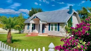 Une maison de plain-pied avec un toit à pignon, un porche à l’avant et un petit jardin devant, agrémenté d’une clôture en bois peinte en blanc et d’un arbuste en fleurs.