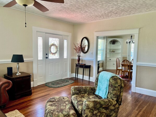 A living room with a patterned armchair, a small table with a lamp, a wooden floor, a door with a wreath, and a dining area in the background. - Charming Sebring Historic Registry Home Near Circle and Across from Lake Jackson (Sebring)