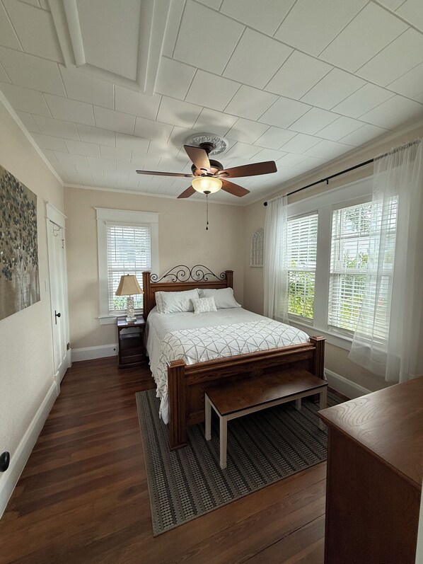 Une chambre avec un lit en bois, un ventilateur de plafond et de grandes fenêtres équipées de stores.