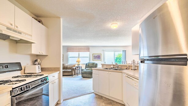 A kitchen with white cabinets, a stainless steel refrigerator, a stove, and a dishwasher. There is a window with blinds and a chair in the background. - Sunny Home in The Villages w/ Lanai & Pool Access! (The Villages)