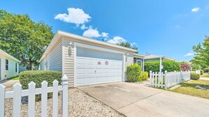 A single-story house with a white garage door, a picket fence, and a gravel driveway. - Sunny Home in The Villages w/ Lanai & Pool Access! (The Villages)
