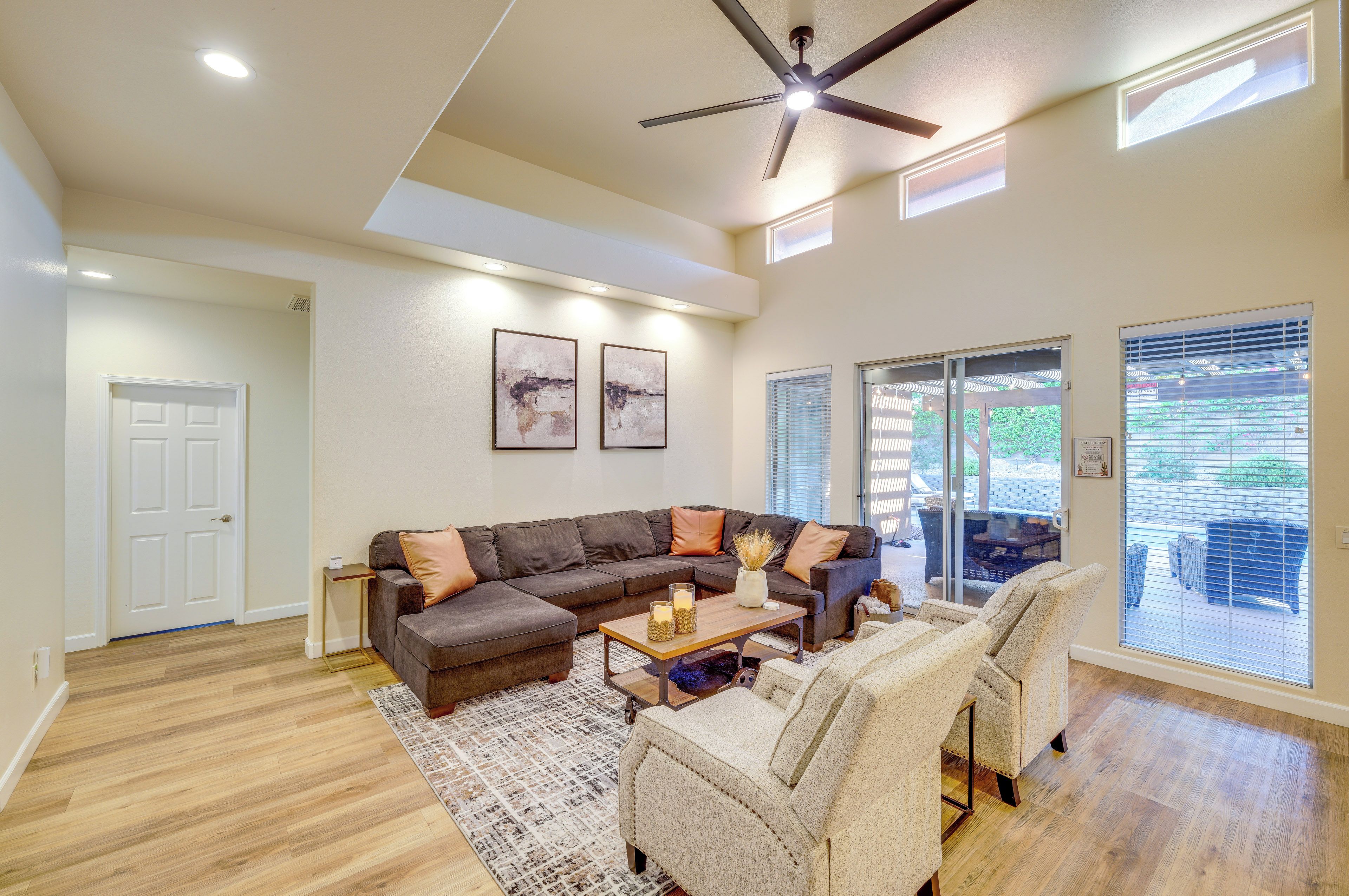 A spacious living room with a sectional sofa, a coffee table, and a fireplace. There is a ceiling fan and a large window providing natural light.