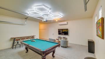 A pool table with a hexagonal ceiling light fixture, a television mounted on the wall, and a painting on the wall.