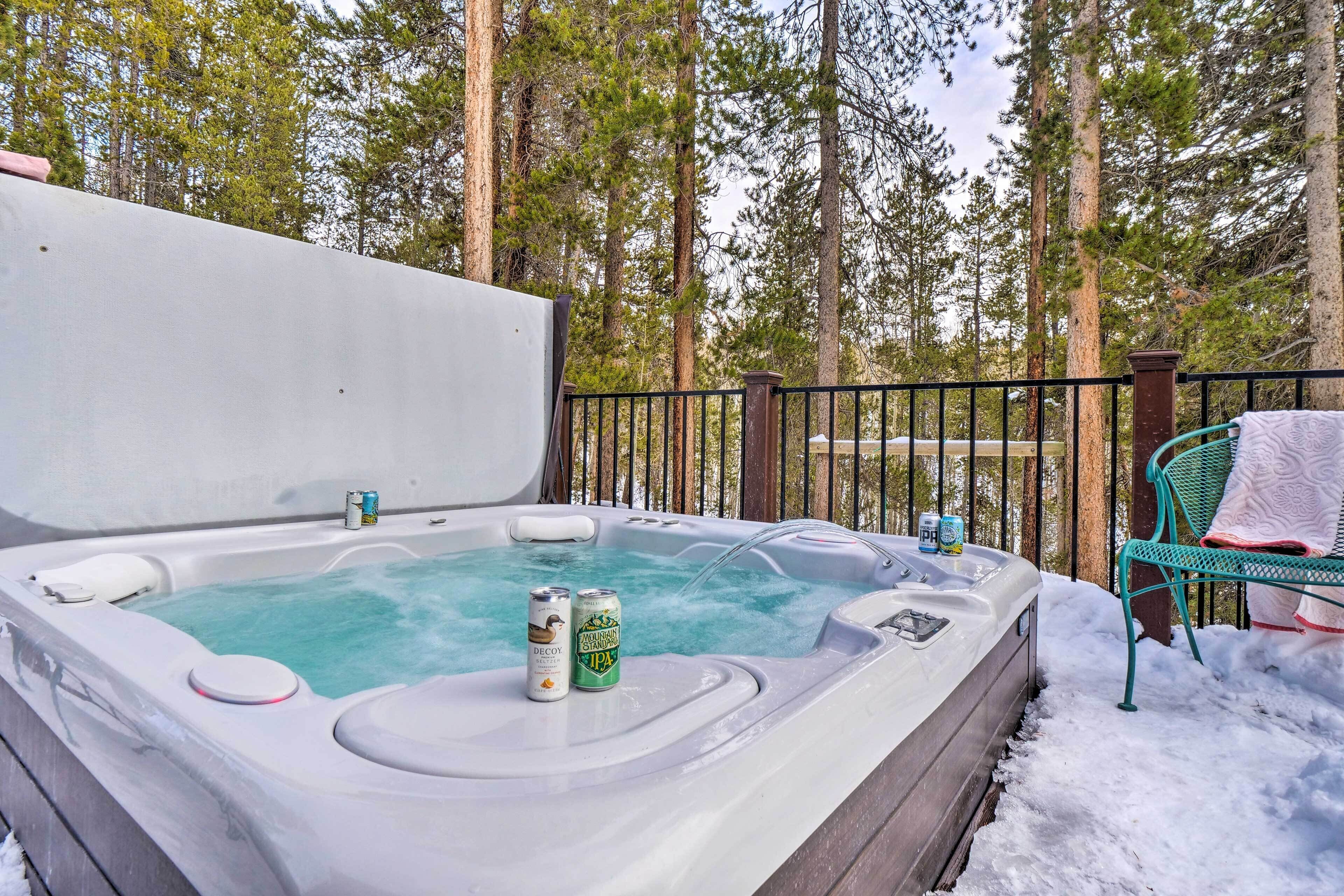 Un bain à remous avec deux canettes de bière, entouré d’une terrasse en bois et de neige, avec de grands arbres en arrière-plan.