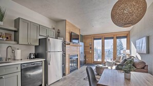 A modern kitchen with grey cabinets, a dishwasher, and a refrigerator. There is a dining area with a wooden table and chairs, and a large window offering a view of the outdoors.