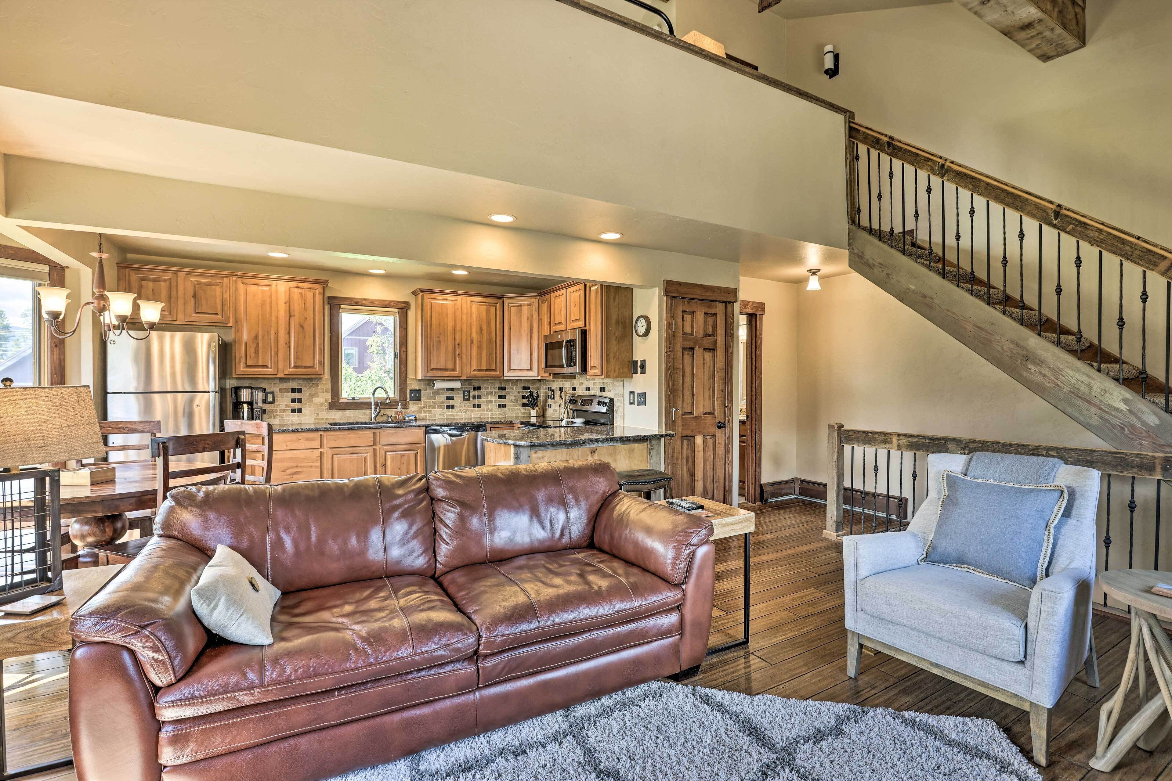 A spacious living room with a leather sofa and a light-colored armchair, a kitchen with wooden cabinets, and a wooden staircase.