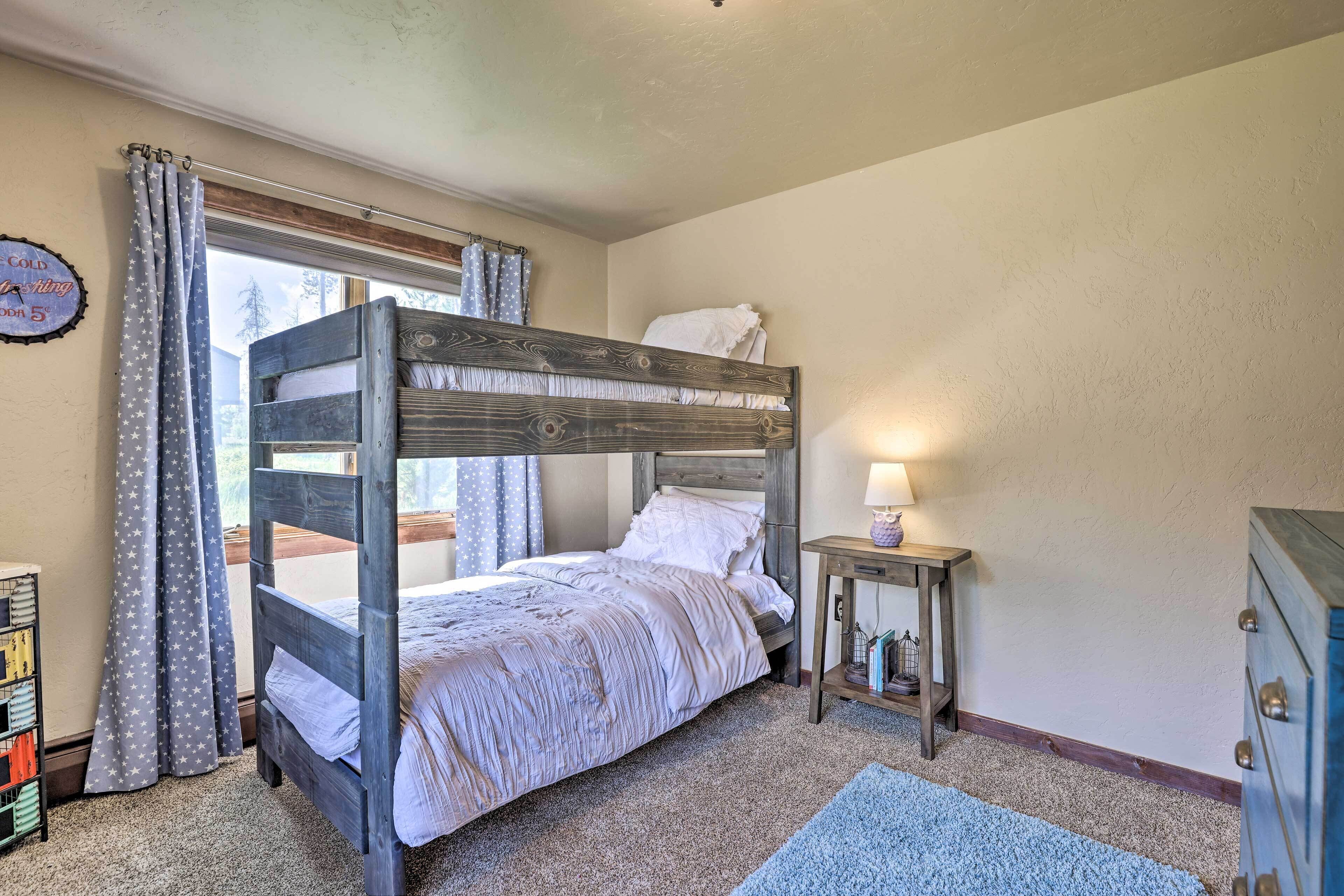 Una litera con cabecera de madera, una mesita de noche con lámpara, una pequeña estantería y una ventana con cortinas.