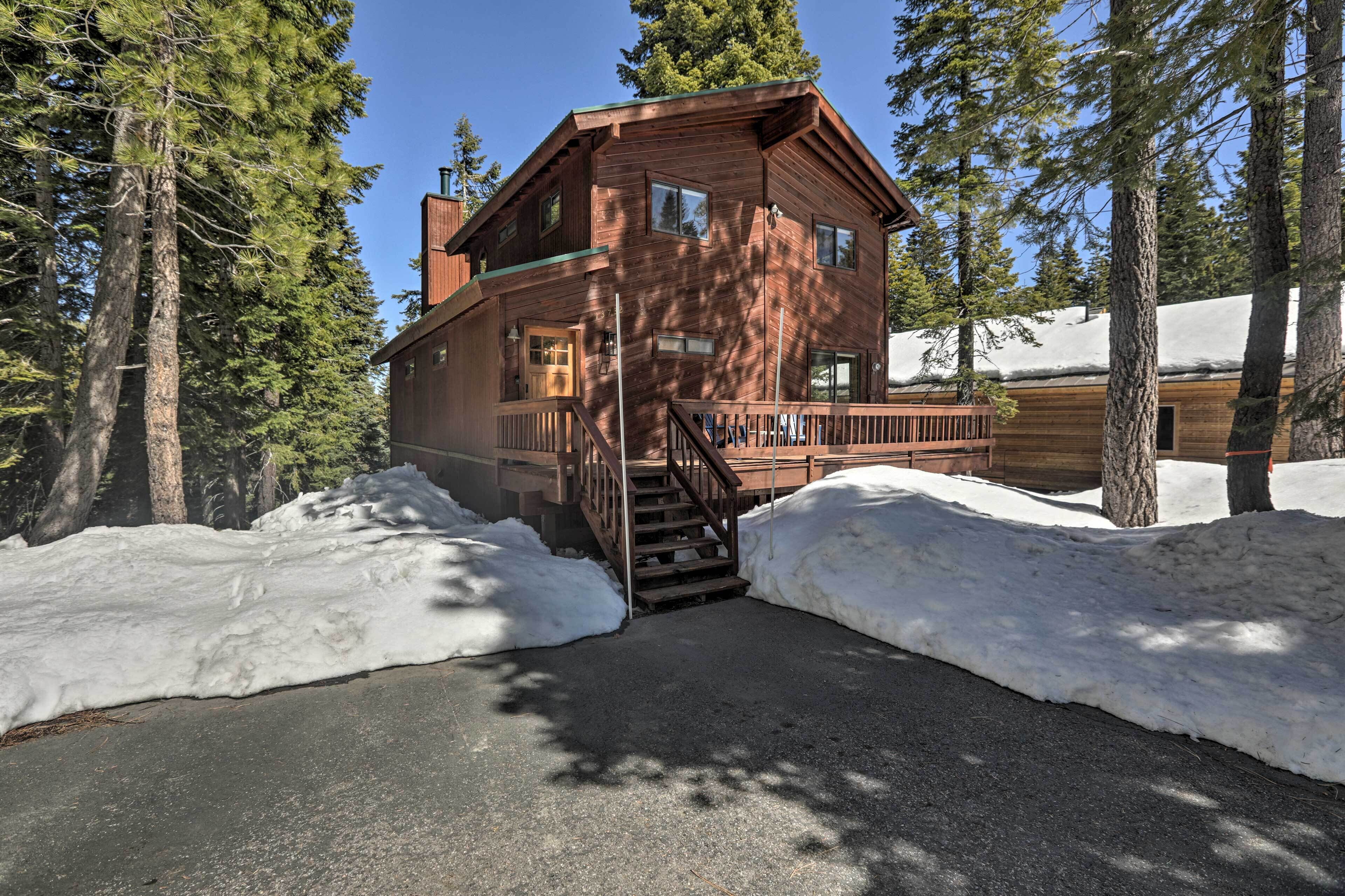 A wooden cabin with a porch and stairs leading to the entrance, surrounded by snow and tall trees.