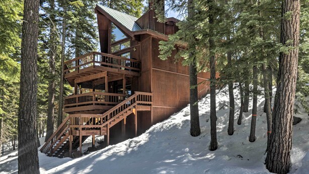 A wooden cabin with a staircase leading to a deck, surrounded by tall trees and snow.