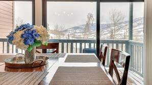A dining table with placemats, a vase with flowers, and a view of a snowy landscape through large windows. - Ski, Shred & Tube: Mtn-view Condo in Silverthrone! (Silverthorne)