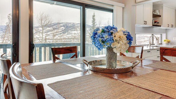 A dining area with a large window, a vase of flowers, and a kitchen in the background. - Ski, Shred & Tube: Mtn-view Condo in Silverthrone! (Silverthorne)