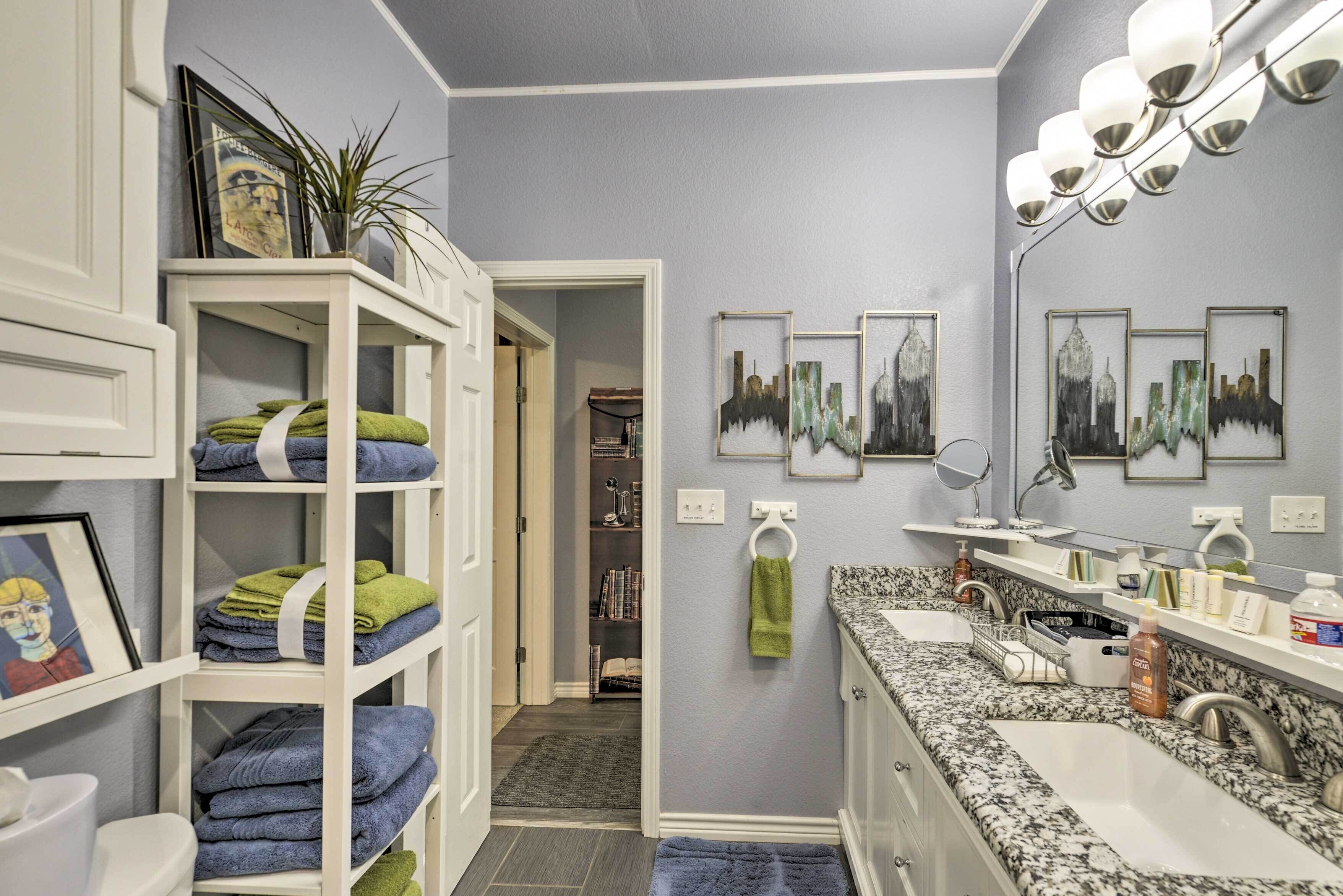 A bathroom with a double sink, a towel rack, and a mirror. There are shelves with folded towels and a framed picture on the wall.
