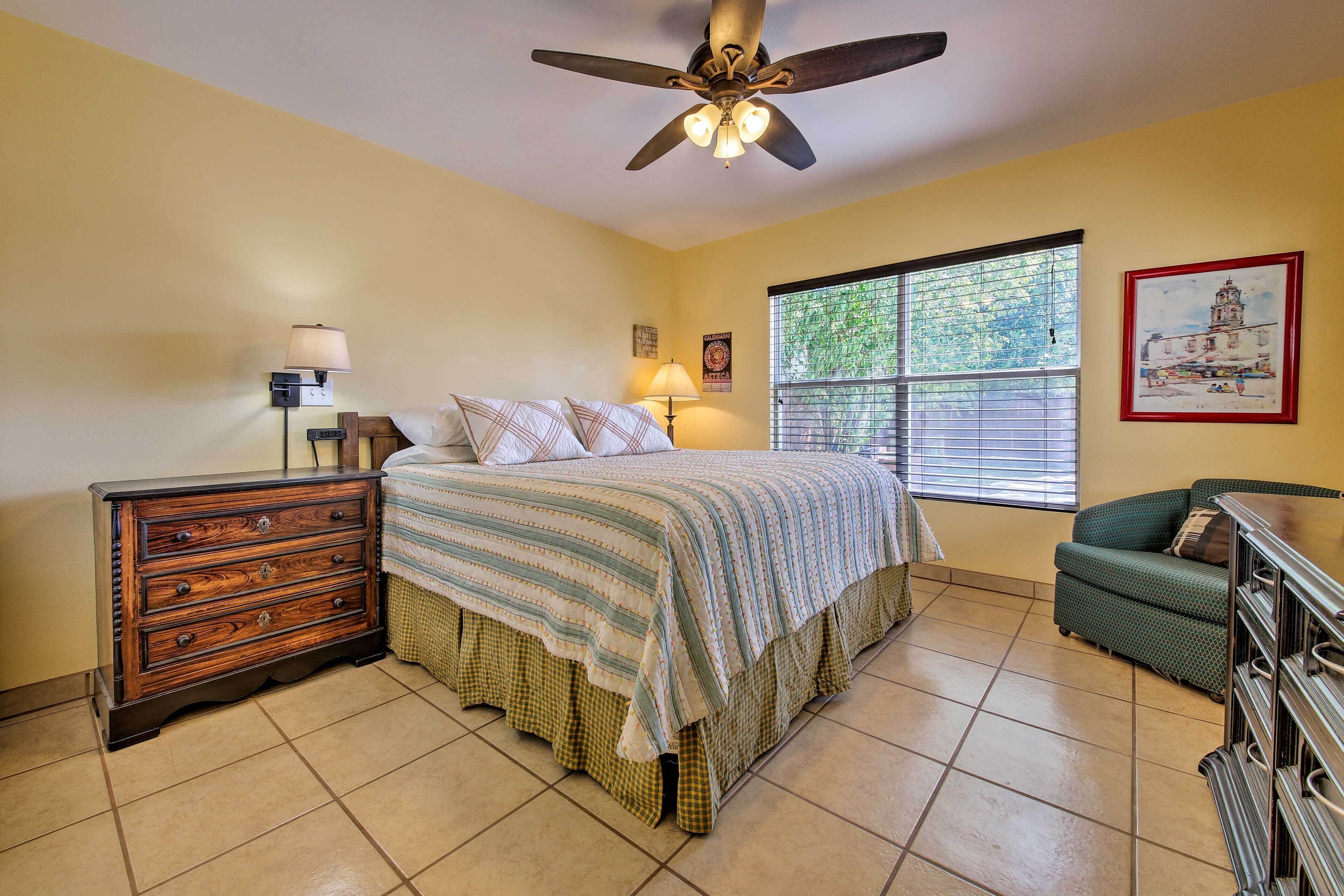 A bedroom with a bed, dresser, lamp, ceiling fan, window, and artwork on the wall.
