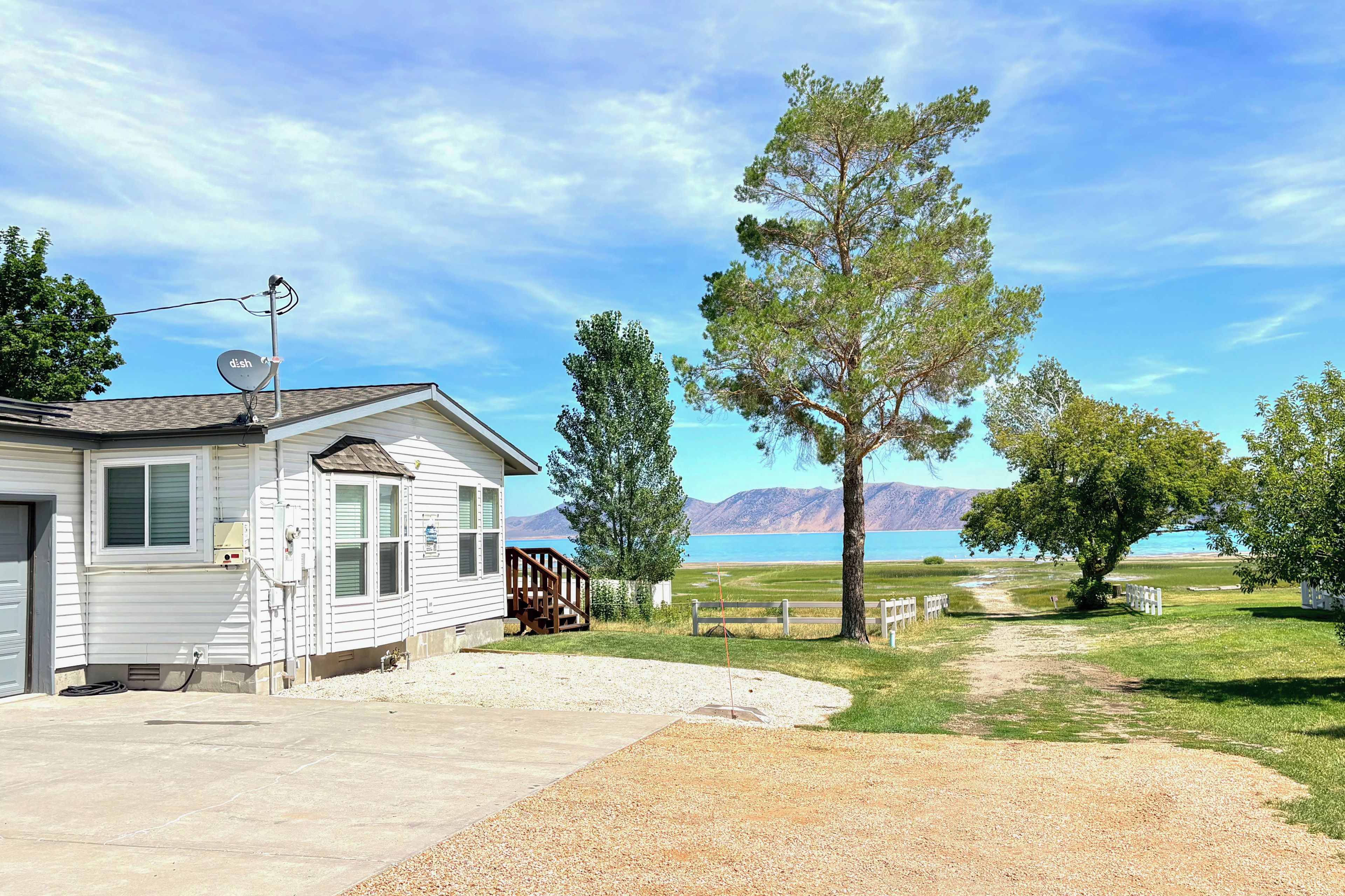 Une maison mobile de plain-pied, dotée d’une antenne parabolique, d’une allée en gravier et offrant une vue sur un lac, avec des montagnes en arrière-plan.